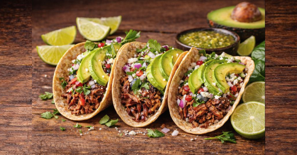 Three healthy brisket tacos made with slow cooker shredded beef, avocado slices, cilantro, red onion, cotija cheese, and salsa verde on a wooden table.